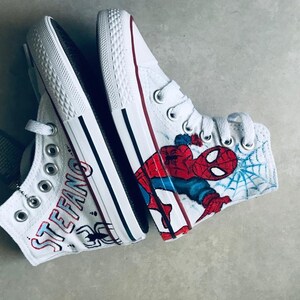 May include: A pair of white high-top sneakers with custom designs. One shoe features a Spiderman graphic with a blue and red color scheme. The other shoe has the name "STEFANO" written in black and red lettering.
