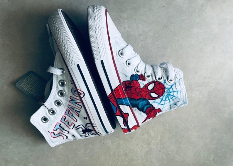May include: A pair of white high-top sneakers with custom designs. One shoe features a Spiderman graphic with a blue and red color scheme. The other shoe has the name "STEFANO" written in black and red lettering.