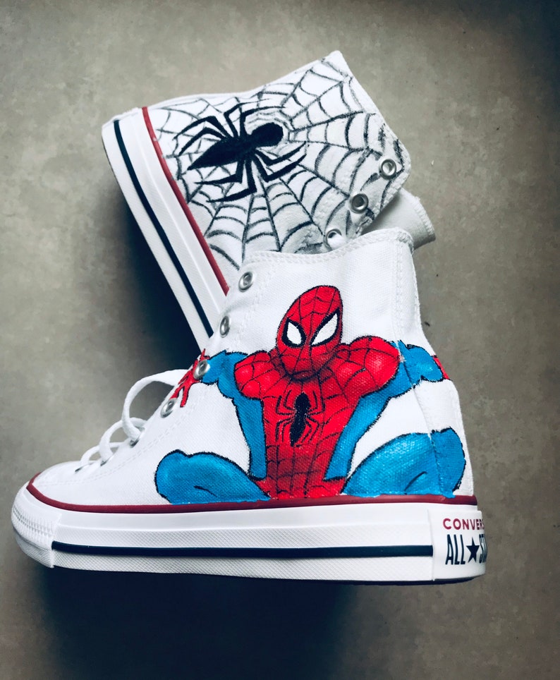 May include: White high-top sneakers with a hand-painted design of Spider-Man on one shoe and a spider web with a spider on the other shoe. The shoes have black laces and a black stripe around the sole. The text "CONVERSE ALL STAR" is visible on the side of the shoe.