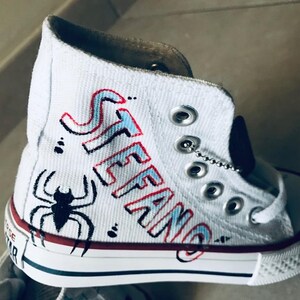 May include: White high-top canvas sneakers with red and blue accents. The name "STEFANO" is written in a stylized font on the side of the shoe. A black spider is drawn on the toe of the shoe.