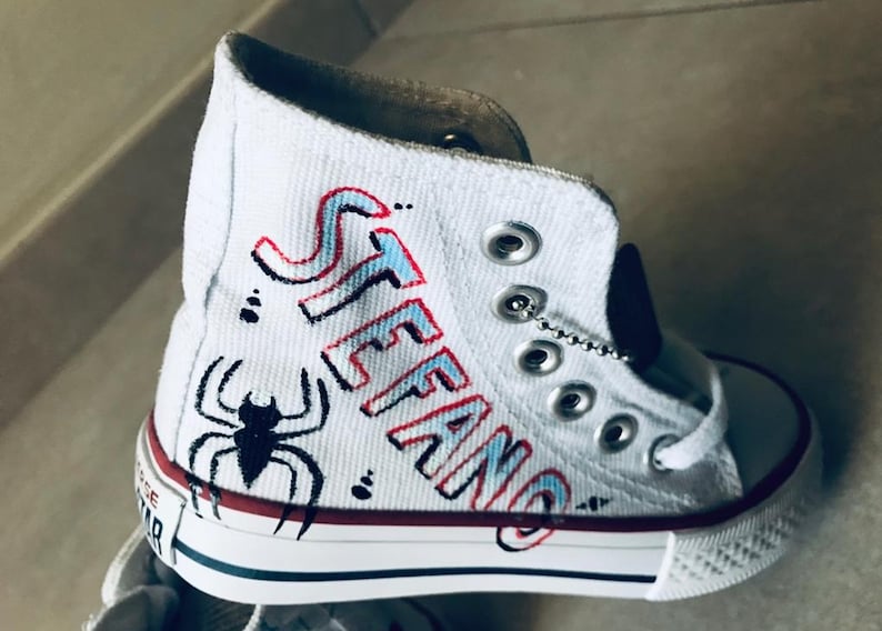 May include: White high-top canvas sneakers with red and blue accents. The name "STEFANO" is written in a stylized font on the side of the shoe. A black spider is drawn on the toe of the shoe.