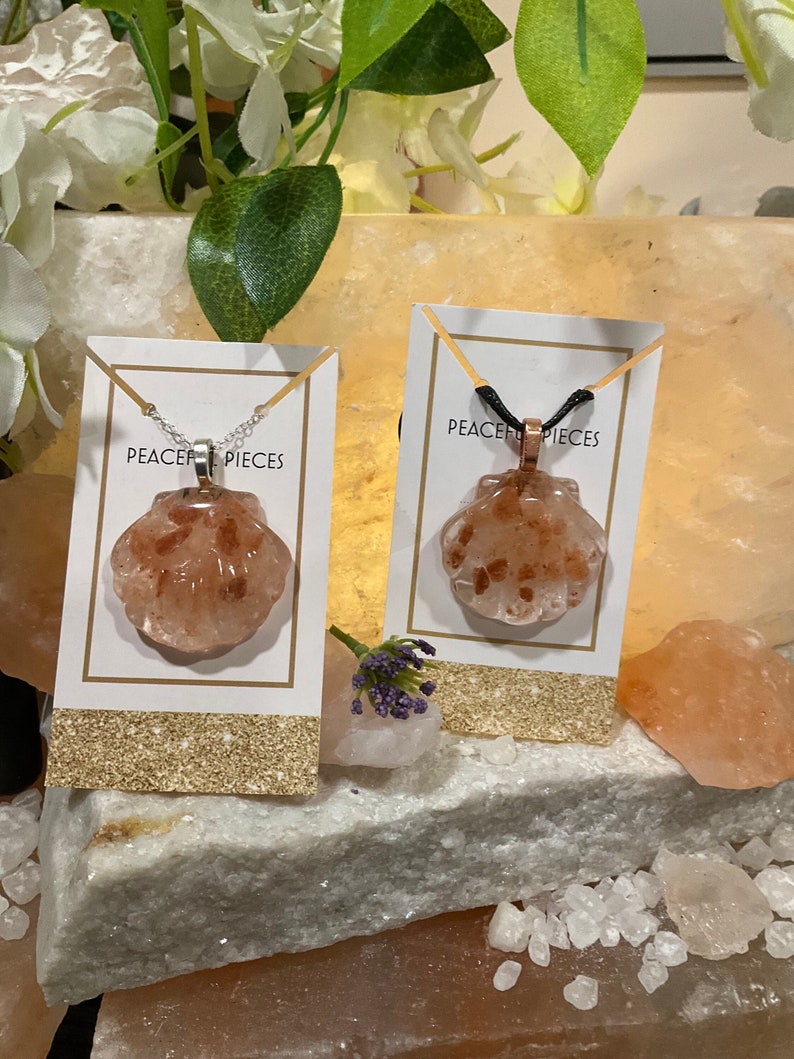 May include: Two necklaces with a seashell pendant made of pink Himalayan salt. The necklaces are displayed on a white card with the text "Peace Pieces" printed on it. The cards are sitting on a bed of pink Himalayan salt crystals.