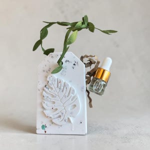 Puede incluir: Una etiqueta rectangular blanca con un patrón de terrazo moteado y un diseño de hoja de monstera en relieve. Un pequeño frasco cuentagotas de vidrio con cuello dorado y tapa blanca está unido al lateral, acentuado por una enredadera de tela.