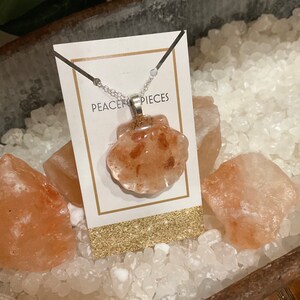 May include: A silver chain necklace with a pink Himalayan salt crystal pendant in the shape of a seashell. The pendant is on a white card with the text "Peace Pieces".