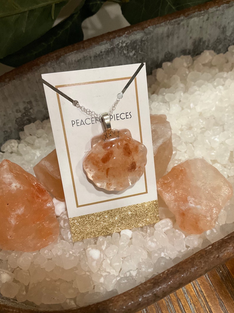 May include: A silver chain necklace with a pink Himalayan salt crystal pendant in the shape of a seashell. The pendant is on a white card with the text "Peace Pieces".