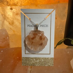 May include: A silver chain necklace with a pink and white seashell pendant. The pendant is made of resin and has a glittery finish. The necklace is displayed on a white card with the text "Peaceful Pieces".