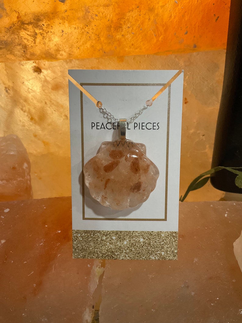 May include: A silver chain necklace with a pink and white seashell pendant. The pendant is made of resin and has a glittery finish. The necklace is displayed on a white card with the text "Peaceful Pieces".