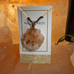 May include: A necklace with a clear, shell-shaped pendant filled with small, orange-brown crystals. The pendant is attached to a black cord and hangs from a gold-colored bail. The necklace is displayed on a white card with the text "PEACE PIECES".