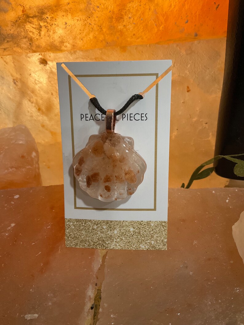 May include: A necklace with a clear, shell-shaped pendant filled with small, orange-brown crystals. The pendant is attached to a black cord and hangs from a gold-colored bail. The necklace is displayed on a white card with the text "PEACE PIECES".