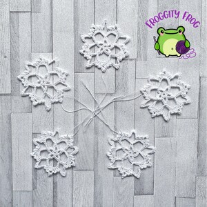 May include: Five white crocheted snowflakes on a woodgrain background. The snowflakes are connected by white yarn.
