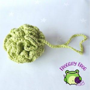 May include: A light green crocheted bath pouf with a loop handle. The pouf is round and textured, with a knotted appearance. The image includes the text "FROGGITY FROG" and a cartoon frog holding a crochet hook and yarn.