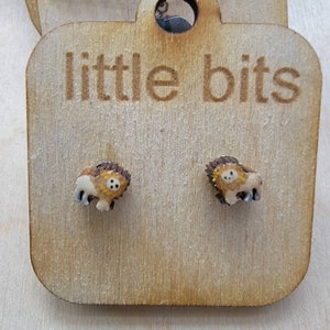 May include: A pair of small, lion-shaped stud earrings. The earrings are tan, brown, and yellow, with a detailed design. They are displayed on a wooden tag with the words "little bits" engraved on it.