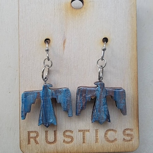 May include: A pair of blue and brown wooden earrings shaped like a stylized bird with silver hooks. The earrings are displayed on a wooden tag with the word "RUSTICS" printed on it.