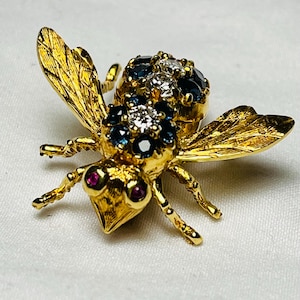 Puede incluir: Un broche de abeja de oro con piedras preciosas azules y blancas. La abeja tiene ojos de rubí y alas detalladas.