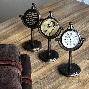 May include: Three antique-style compass stands with a photo and a clock face. The photo compass stand has a black and white photo of a couple. The clock face compass stand has roman numerals and black hands. The third compass stand has a personalized message engraved on the face.  "I will find you in every lifetime, just as I found you in this one. Love you forever and always Sofce"