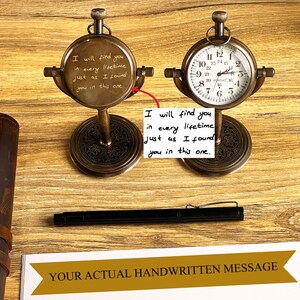 May include: Two antique brass-colored pocket watch clocks on stands. One clock face displays a handwritten message: "I will find you in every lifetime just as I found you in this one." The other clock shows the time. A black pen rests nearby.