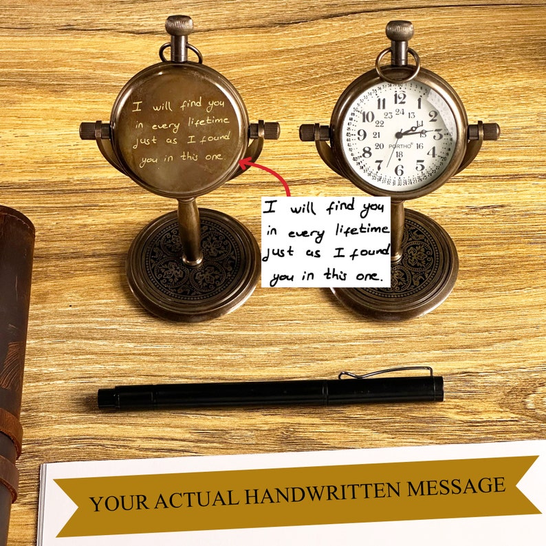 May include: Two antique brass-colored pocket watch clocks on stands. One clock face displays a handwritten message: "I will find you in every lifetime just as I found you in this one." The other clock shows the time. A black pen rests nearby.