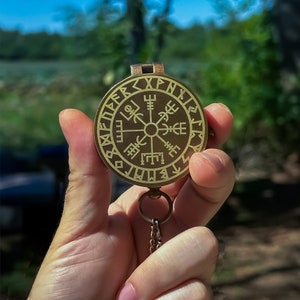 May include: A brass compass with a chain, featuring a circular design with runes and symbols. The compass is held in a hand.