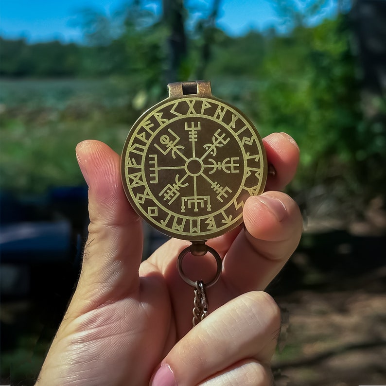 May include: A brass compass with a chain, featuring a circular design with runes and symbols. The compass is held in a hand.