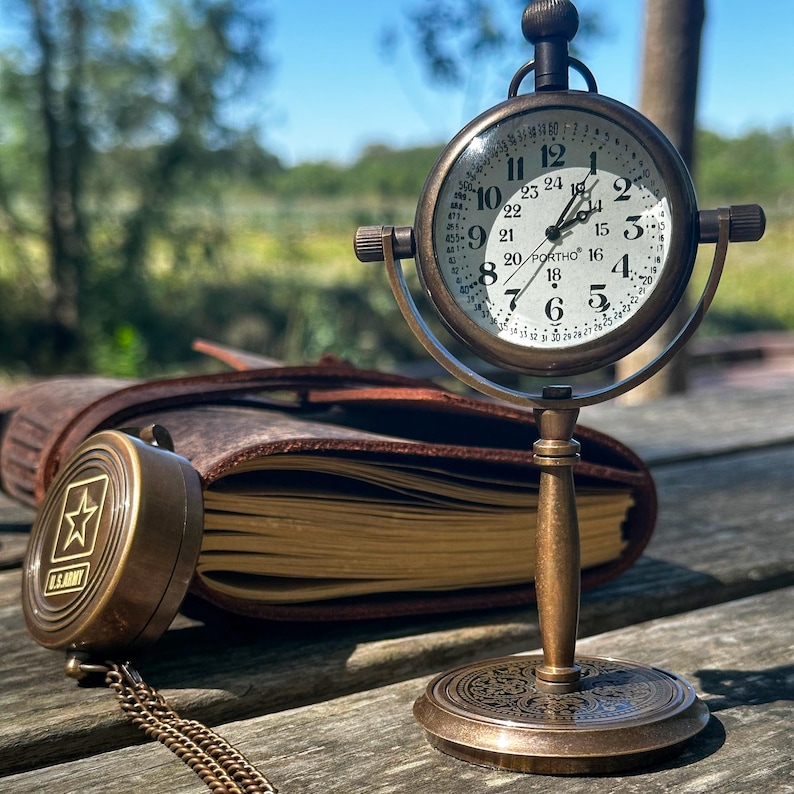 K&ouml;nnte beinhalten: Eine antike Messing-Schreibtischuhr mit rundem Zifferblatt und r&ouml;mischen Ziffern. Die Uhr wird von einem dekorativen St&auml;nder getragen. Ein U.S. Army Kompass und ein ledergebundenes Journal sind ebenfalls zu sehen.