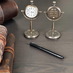 May include: Two antique-style desk clocks with a silver finish, one displaying a clock face and the other engraved with text. A black pen and leather-bound journals are also visible on the dark wooden surface.