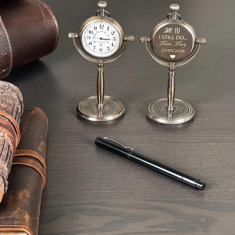 May include: Two antique-style desk clocks with a silver finish, one displaying a clock face and the other engraved with text. A black pen and leather-bound journals are also visible on the dark wooden surface.