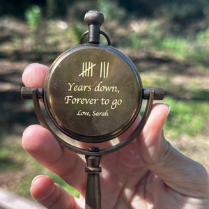 May include: A vintage brass compass with the inscription "Years down, Forever to go, Love, Sarah." The compass face features tally marks and is held by a curved handle. The compass is a warm, antique gold color.