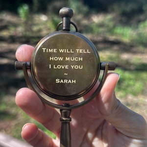 K&ouml;nnte beinhalten: Ein antiker Messingkompass mit der Inschrift "TIME WILL TELL HOW MUCH I LOVE YOU ~ SARAH". Der Kompass hat ein kreisf&ouml;rmiges Zifferblatt, einen Knopf und einen Griff. Der Hintergrund ist verschwommenes gr&uuml;nes Laub.