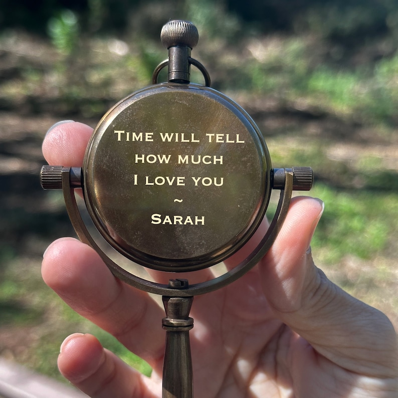 K&ouml;nnte beinhalten: Ein antiker Messingkompass mit der Inschrift "TIME WILL TELL HOW MUCH I LOVE YOU ~ SARAH". Der Kompass hat ein kreisf&ouml;rmiges Zifferblatt, einen Knopf und einen Griff. Der Hintergrund ist verschwommenes gr&uuml;nes Laub.