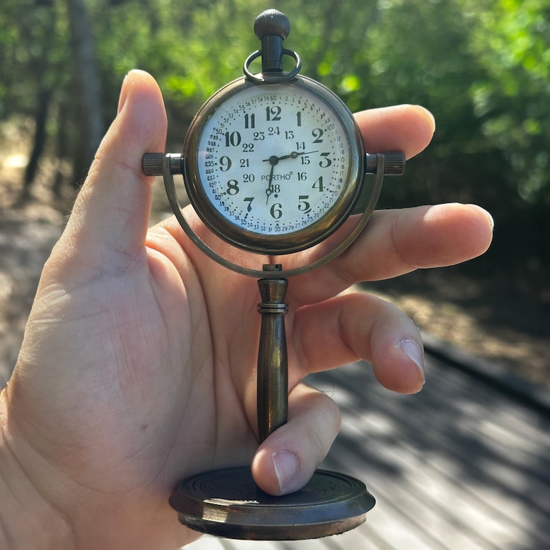 May include: A vintage-style brass desk clock with a white face and black numerals. The clock has a round face with a small handle at the top and is supported by a stand. The clock face displays the time as 2:16.