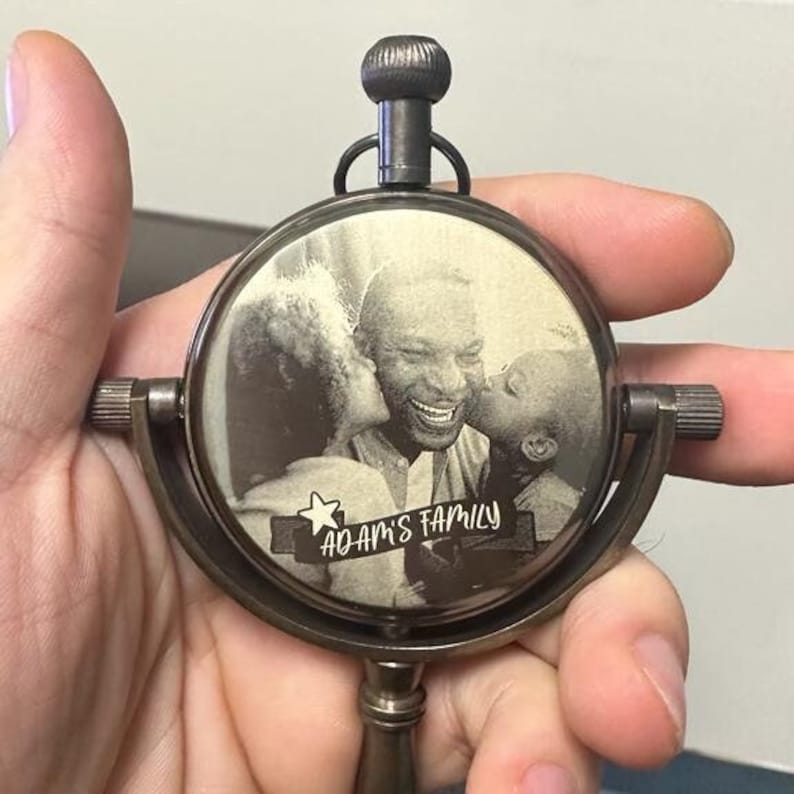 May include: A vintage-style, circular, metal pendant with a family photo inside. The photo is black and white and shows a man being kissed by two people. The words "ADAM'S FAMILY" are etched on a banner below the photo.