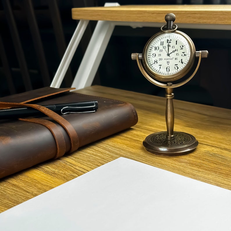 K&ouml;nnte beinhalten: Eine Tischuhr aus Messing im Vintage-Stil mit wei&szlig;em Zifferblatt und schwarzen Ziffern. Die Uhr ist auf einem dekorativen St&auml;nder montiert. Ein braunes Lederjournal mit einem Stift und ein wei&szlig;es Blatt Papier liegen ebenfalls auf dem Holzschreibtisch.