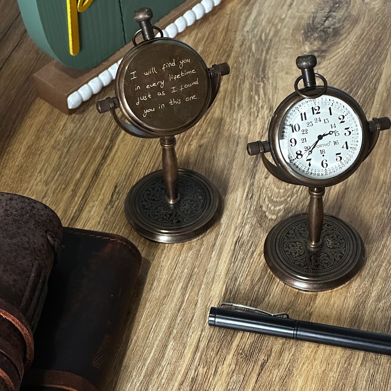 K&ouml;nnte beinhalten: Zwei Schreibtischuhren im antiken Stil mit bronzefarbenen Metallrahmen und -st&auml;ndern. Eine Uhr zeigt ein wei&szlig;es Zifferblatt mit schwarzen Ziffern, die andere handschriftlichen Text. Ein schwarzer Stift und Lederartikel sind ebenfalls sichtbar.