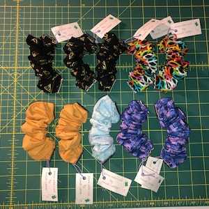 Peut inclure: Huit chouchous colorés avec différents motifs, y compris des paillettes dorées, du tie-dye et des imprimés floraux. Chaque chouchou est accompagné d'une petite étiquette indiquant "Sewn with Love by Donat Larose".