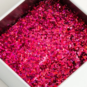 Op de afbeelding: Close-up van een witte vierkante container gevuld met levendige, felroze glitter. De glitter bestaat uit kleine, platte, zeshoekige stukjes, waardoor een sprankelend, getextureerd oppervlak ontstaat. De binnenkant van de container is diep magenta.
