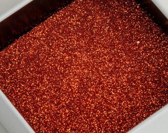 May include: Close-up of a white square container filled with sparkling, reddish-orange glitter. The glitter is densely packed, reflecting light and creating a shimmering effect. The container has a dark brown interior.