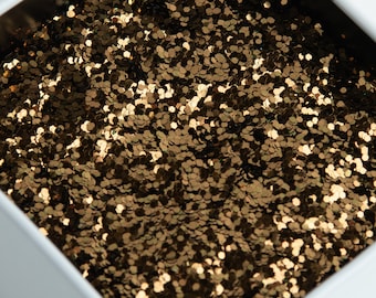 Puede incluir: Primer plano de un recipiente cuadrado blanco lleno de purpurina dorada. La purpurina está formada por pequeñas piezas hexagonales planas que reflejan la luz. El recipiente está sobre un fondo blanco.