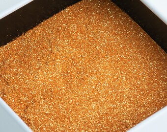 May include: A square white container filled with sparkling gold glitter. The glitter has a fine texture and reflects light, creating a shimmering effect. The container's interior is dark, contrasting with the bright glitter.