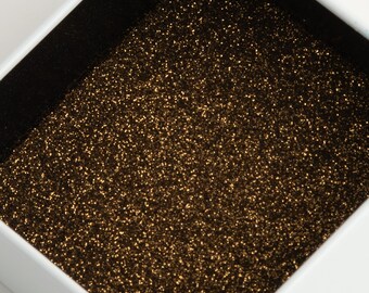 Puede incluir: Una caja de joyería blanca abierta con un interior negro lleno de purpurina dorada. La caja revela un contenido brillante. La purpurina está densamente empaquetada, creando un efecto brillante.