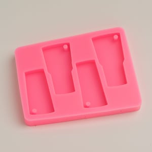 Puede incluir: Un molde de silicona rosa brillante con cuatro cavidades en forma de vaso. Cada cavidad tiene un pequeño agujero en la parte superior para sujetar un llavero. El molde es rectangular con esquinas redondeadas.