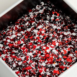 May include: A white square container filled with red, black, and white glitter. The glitter pieces are various shapes and sizes, creating a textured, sparkling effect. The container has a black interior.