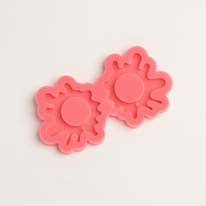 May include: A pair of linked, pink, flower-shaped silicone molds. Each mold has a central circle and a wavy, irregular outer edge. The molds are designed for crafting or creating decorative items.