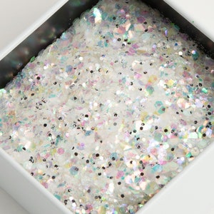 May include: Close-up of a white square container filled with iridescent glitter. The glitter reflects multiple colors, including pink, blue, and green. The container has a black interior and white exterior.
