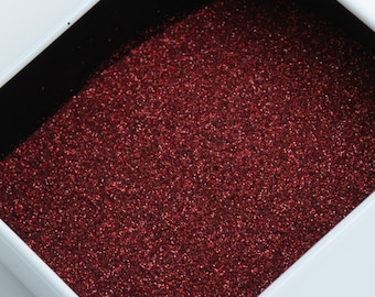 May include: Close-up of a white square box filled with sparkling red glitter. The glitter is densely packed, creating a textured surface. The box has a black interior, contrasting with the bright glitter.