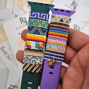 以下が含まれることがあります： カラフルなビーズのデザインが施されたApple Watchバンド2本。左側のバンドは緑色で、さまざまなカラフルなビーズパターンが施されています。右側のバンドは紫色で、紫、ピンク、オレンジ、黄色のストライプのビーズパターンが施されています。