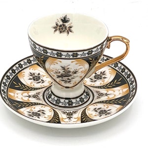 May include: A white teacup and saucer set with a black and gold floral design. The cup has a wide, flared base and a delicate handle. The saucer has a matching design and a scalloped edge.