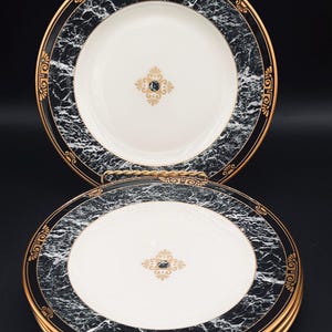 May include: Set of four white dinner plates with a black and gold marble design. The plates have a gold trim and a gold design in the center.