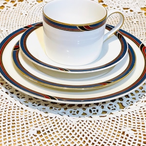 May include: A white teacup and saucer set with a gold rim and a navy blue, red, and gold design. The set includes a teacup, a saucer, and a small plate. The set is on a white lace tablecloth.