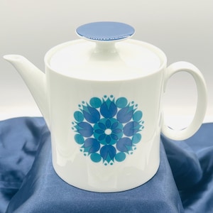 May include: A white ceramic teapot featuring a circular blue and teal floral-inspired geometric pattern on the front. The teapot has a matching white lid topped with a flat, circular blue knob, a curved handle, and a tapered spout.