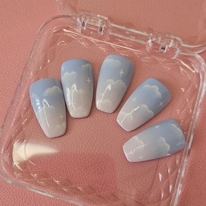 Könnte beinhalten: Fünf künstliche Fingernägel mit einem Himmelsmotiv. Jeder Nagel hat eine hellblaue Basis mit weißen Wolken- und Sternakzenten. Die Nägel werden in einer transparenten, offenen Plastikbox auf rosa Hintergrund präsentiert.